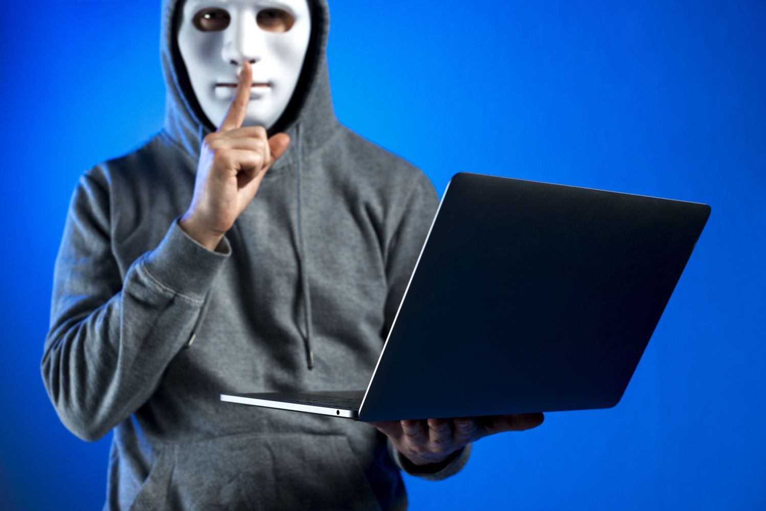 Person in a gray hoodie wearing a white mask holds a laptop and shushes with a finger to lips suggesting a hacker or secretive activity