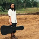 Man with long hair and glasses stands on a dirt construction site holding a black guitar case