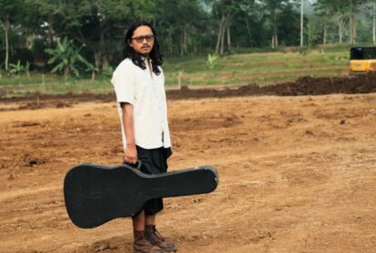Man with long hair and glasses stands on a dirt construction site, holding a black guitar case.