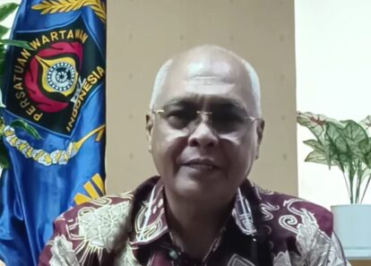 Close-up of an older man wearing glasses speaking in a room, with a blue organizational flag in the background.