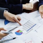 Business professionals compare financial charts and graphs spread on a table using a calculator and pointing at data sheets