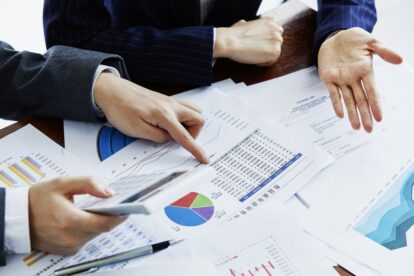 Business professionals compare financial charts and graphs spread on a table, using a calculator and pointing at data sheets.