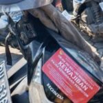 Red poster with the word kewajiban attached to a black honda scooter parked among other motorcycles on a stone paved surface
