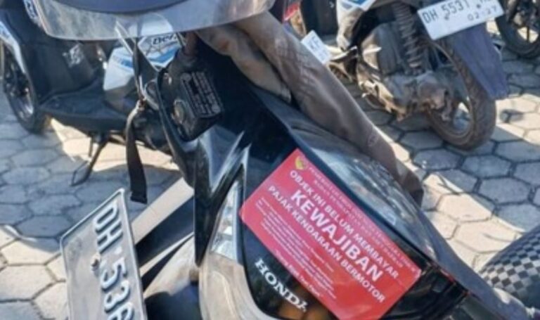 Red poster with the word KEWAJIBAN attached to a black Honda scooter parked among other motorcycles on a stone-paved surface.