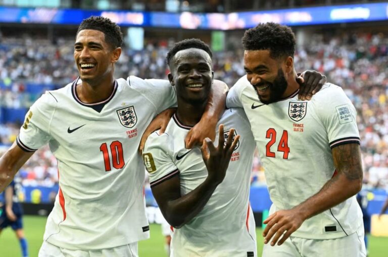 Three England players celebrate together on the pitch, smiling with arms around each other after a goal, stadium cheering in the background.