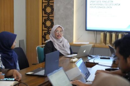 Woman in a light gray hijab speaking during a meeting at a wooden table with laptops; presentation screen visible in the background.