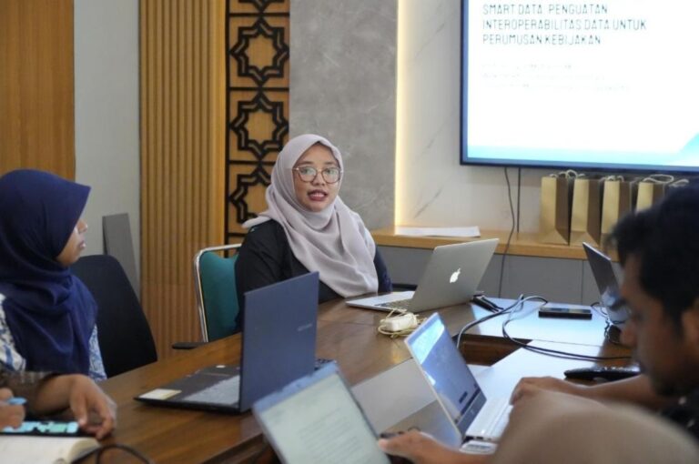 Woman in a light gray hijab speaking during a meeting at a wooden table with laptops; presentation screen visible in the background.
