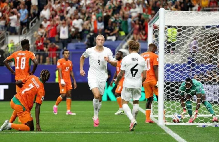 Soccer players in white and orange near the goal, with the ball at the goal line and the goalkeeper on the ground nearby.