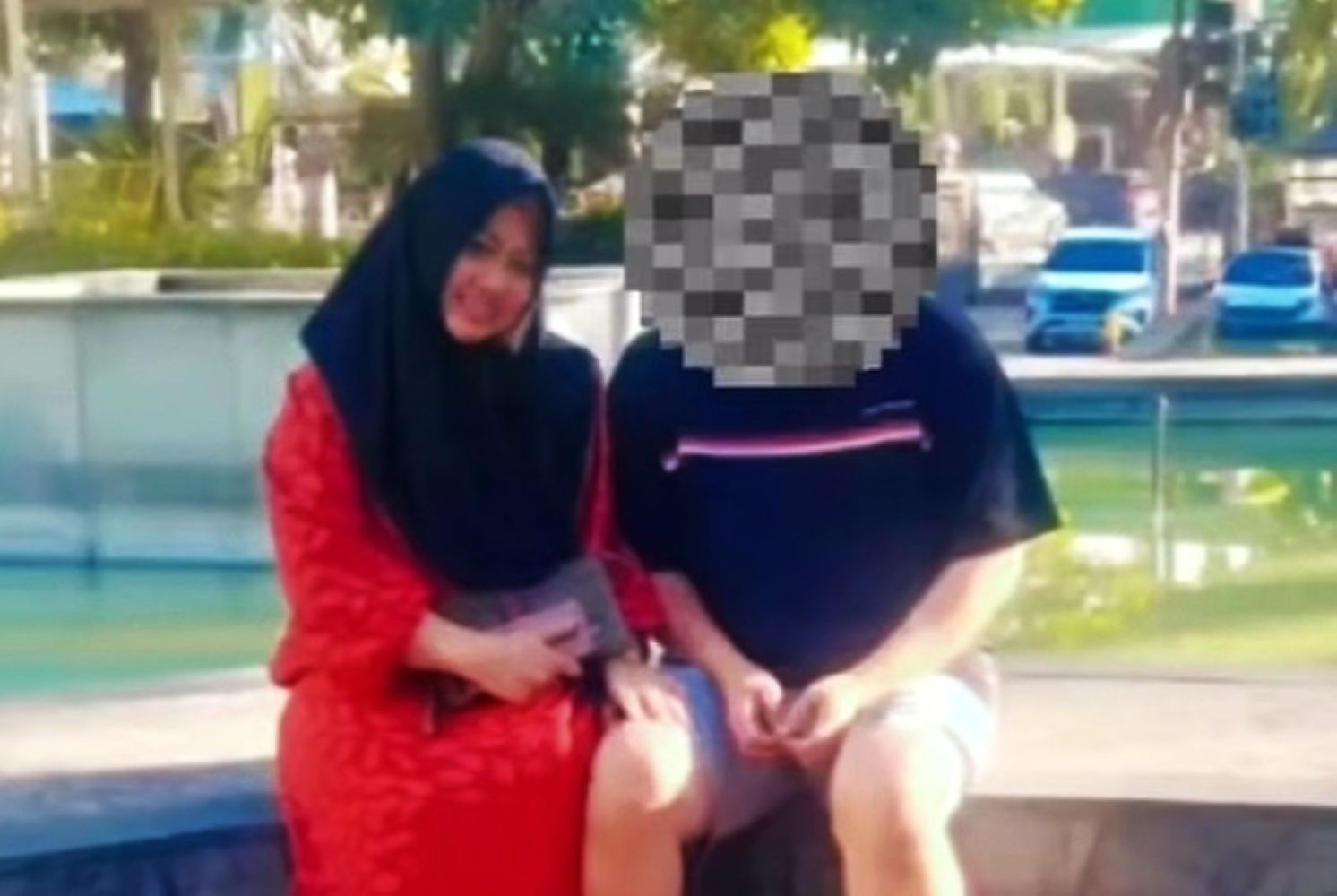 Two people sit on a low wall by a sunny park fountain a woman in a red dress and black hijab sits beside a man in a dark shirt whose face is pixelated or blurred