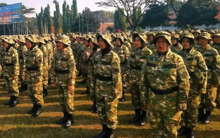 Group of soldiers in camouflage uniforms standing in formation on a grassy field on a sunny day.