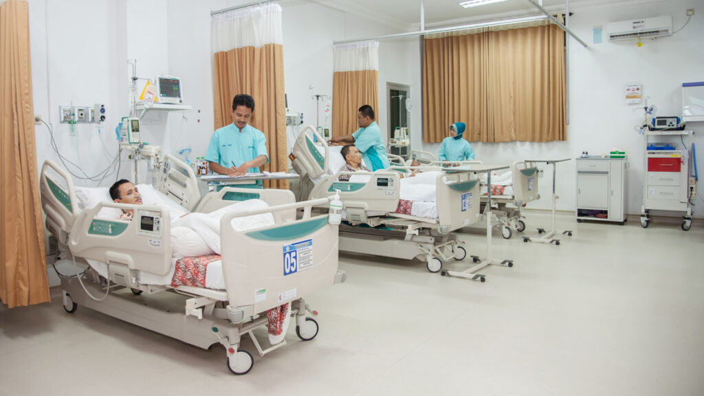 Hospital ward with multiple patients in adjustable beds nurses in teal scrubs attending to them