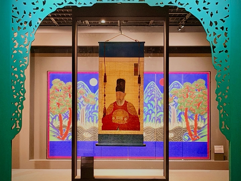 20260707111620375474wcyd0 catrawarta Museum display of a hanging scroll portrait of a seated scholar in red robes framed by a glass case and teal decorative arch with vibrant background artwork behind