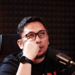 Man wearing glasses and a black shirt sits at a microphone in a recording studio