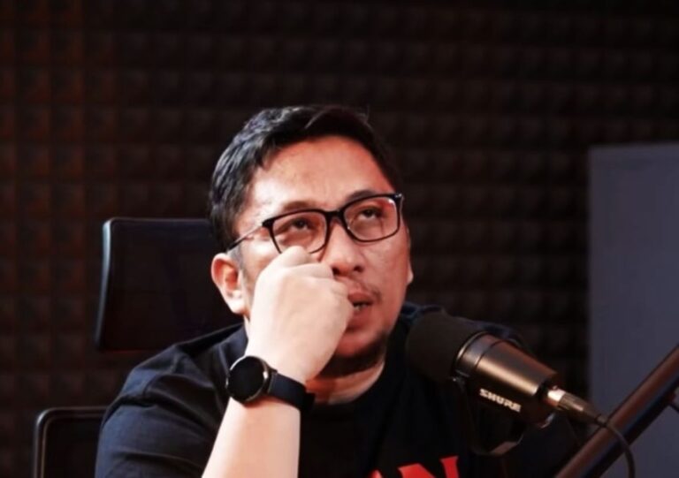 Man wearing glasses and a black shirt sits at a microphone in a recording studio.