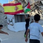 Group of boys walking amid rubble holding spanish and palestinian flags near a white tent
