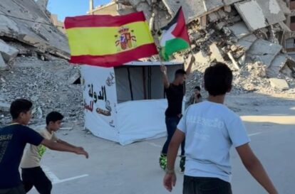 Group of boys walking amid rubble, holding Spanish and Palestinian flags near a white tent.