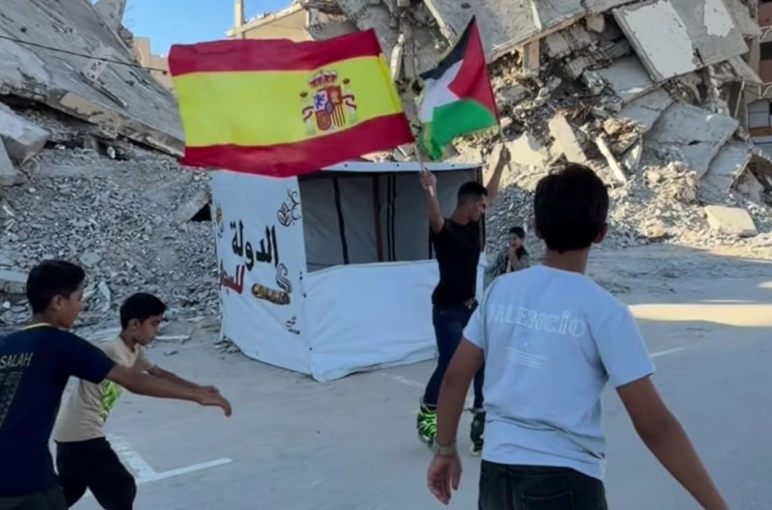 20ggaza catrawarta Group of boys walking amid rubble holding spanish and palestinian flags near a white tent