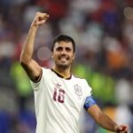Male soccer player in a white jersey with number 16 raises a clenched fist in celebration on the field crowd in the background