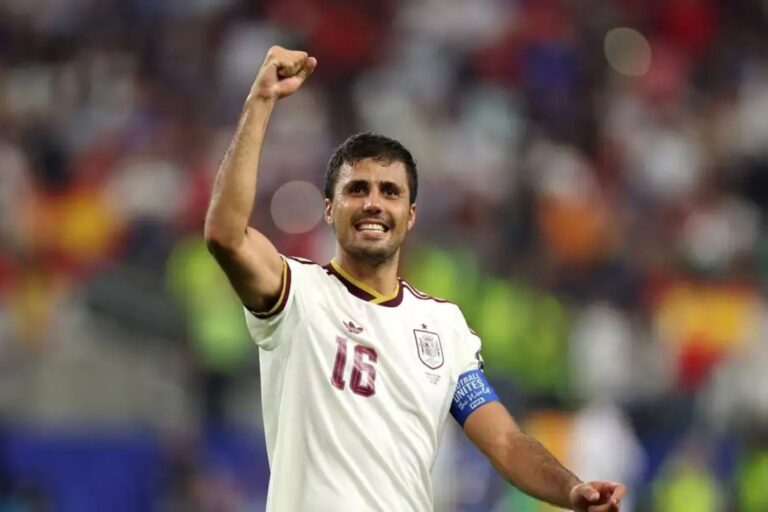 Male soccer player in a white jersey with number 16 raises a clenched fist in celebration on the field, crowd in the background.
