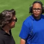 Two people on a grassy field talk man in blue athletic shirt wearing a headset speaks to another person in black