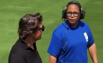 Two people on a grassy field talk; man in blue athletic shirt wearing a headset speaks to another person in black.