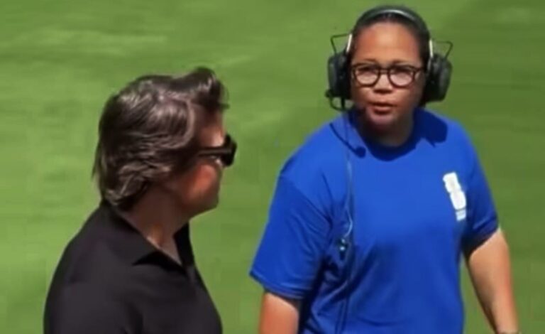 Two people on a grassy field talk; man in blue athletic shirt wearing a headset speaks to another person in black.