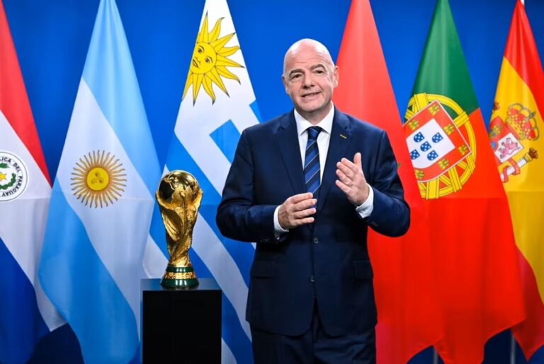 Man in a dark suit stands beside a golden trophy on a pedestal, with multiple national flags in the background.