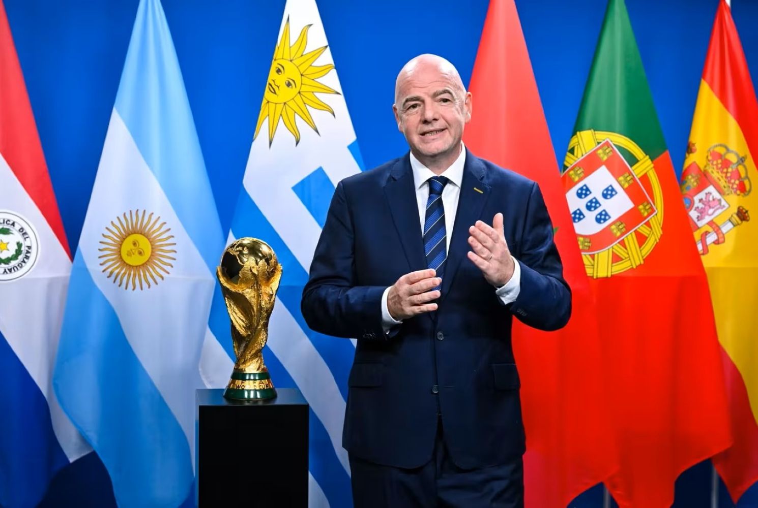 21gfifa1 catrawarta Man in a dark suit stands beside a golden trophy on a pedestal with multiple national flags in the background