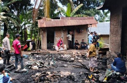 Villagers gather around charred rubble outside a small brick house with a red corrugated roof in a tropical setting.