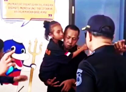 A man in black is held by a young girl as two police officers stand nearby with a cartoon penguin and trident mural in the background