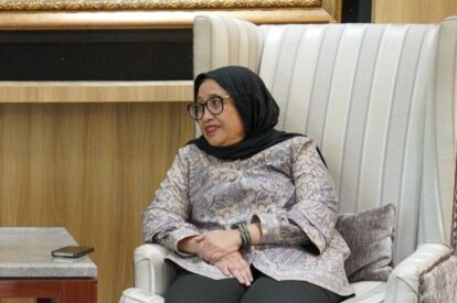 Woman wearing a black hijab and glasses sits in a striped white chair, hands folded on her lap in a formal indoor setting.
