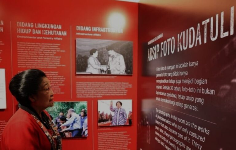 Woman in a red jacket stands at a red exhibition wall reading panels and viewing photos labeled in Indonesian.