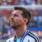 Soccer player with short dark hair and stubble wearing a light blue jersey and a medal on a blue lanyard standing on a pitch
