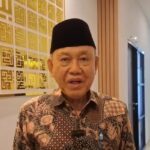 Older man wearing a black cap and batik shirt speaks in a bright hallway a lapel mic clipped to his shirt