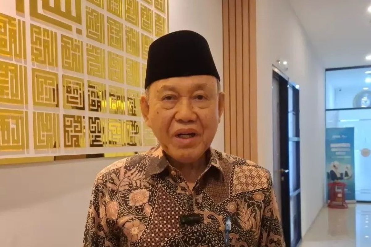 Older man wearing a black cap and batik shirt speaks in a bright hallway a lapel mic clipped to his shirt