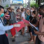 Uniformed officer wearing a mask hands pink documents to people lined up outdoors