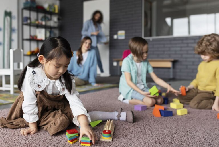 Young girl arranging colorful wooden blocks on a carpet in a classroom while other children play nearby and a teacher stands in the background.