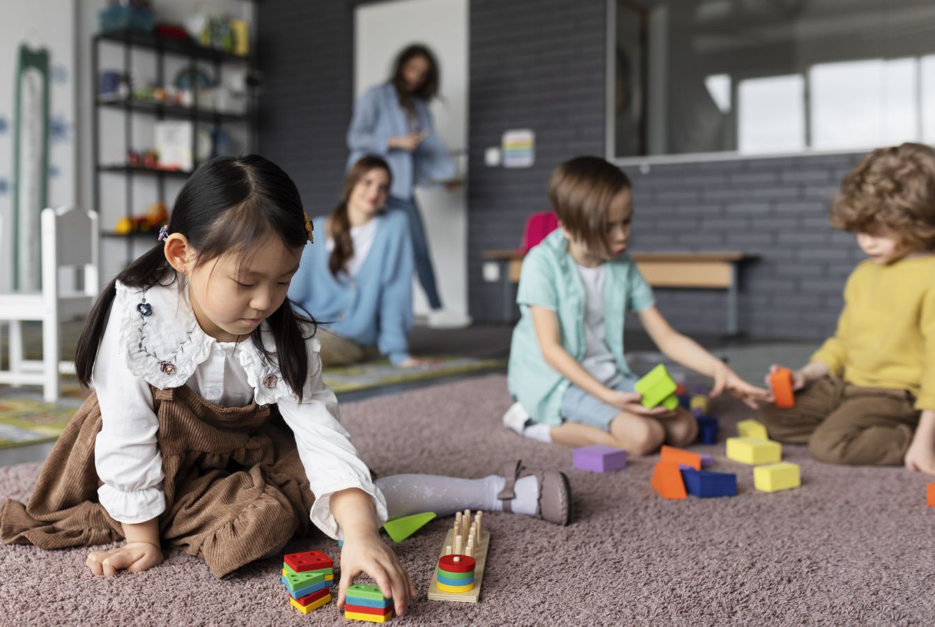 24ganak catrawarta Young girl arranging colorful wooden blocks on a carpet in a classroom while other children play nearby and a teacher stands in the background