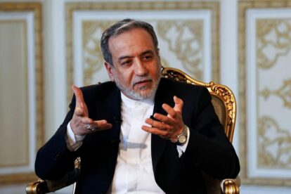 Man in a dark suit sits in an ornate gold chair gesturing with his hands while speaking in an elegant room