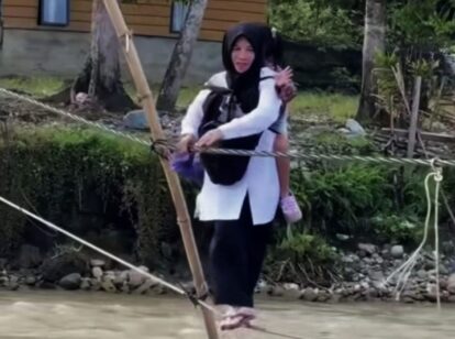 Woman in a black hijab and white tunic carries a child on her back while crossing a rope bridge over a shallow river