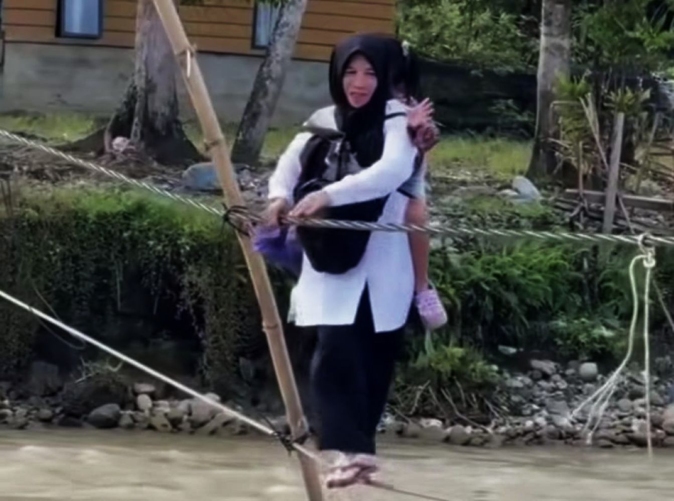 Woman in a black hijab and white tunic carries a child on her back while crossing a rope bridge over a shallow river