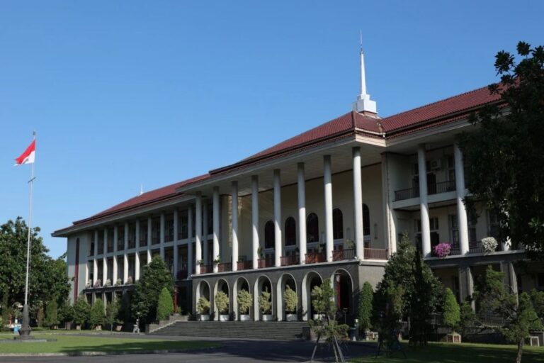 Front view of a grand government building with tall white columns, a red-tiled roof, and an Indonesian flag on a flagpole in front.