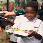 Schoolgirl in a white uniform holds a metal lunch tray with compartments in a crowded outdoor meal line