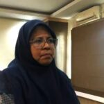 Woman wearing a navy hijab and glasses indoors with a plant and wooden cabinet in the background
