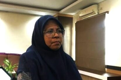 Woman wearing a navy hijab and glasses indoors with a plant and wooden cabinet in the background