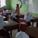 Teacher standing at the front of a small classroom as students sit at wooden desks and listen