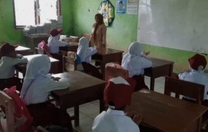 Teacher standing at the front of a small classroom as students sit at wooden desks and listen