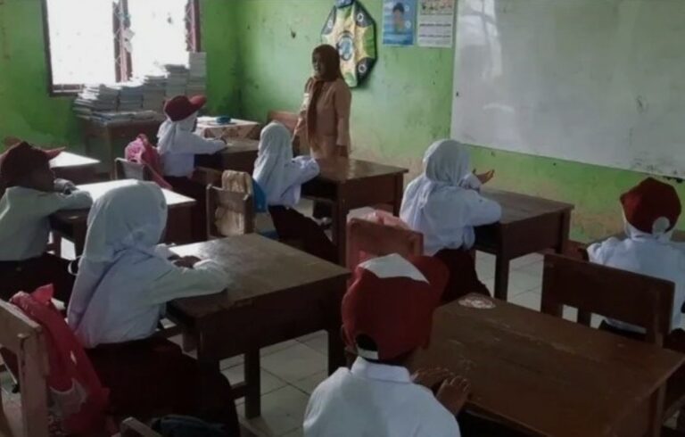 Teacher standing at the front of a small classroom as students sit at wooden desks and listen.