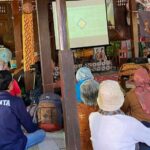 Community workshop in a wooden hall with a speaker presenting slides to attendees seated on the floor in the audience area