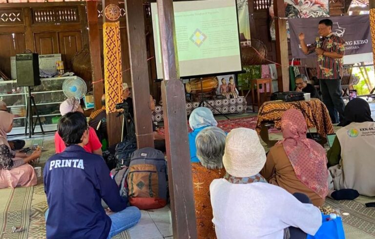 Community workshop in a wooden hall with a speaker presenting slides to attendees seated on the floor in the audience area.
